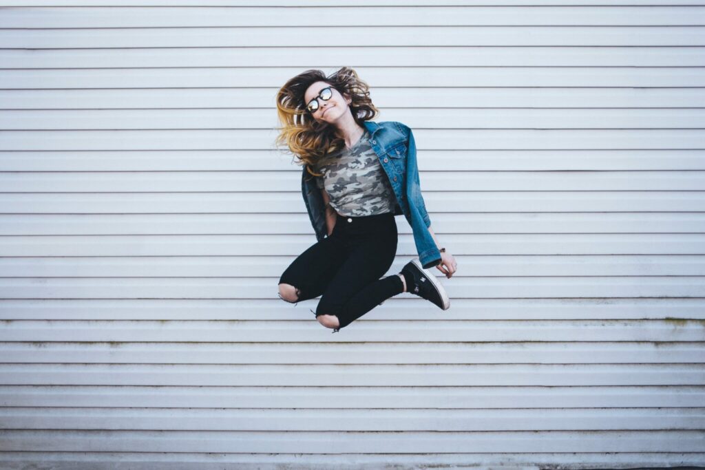 A woman in a denim jacket and glasses joyfully jumps in the air