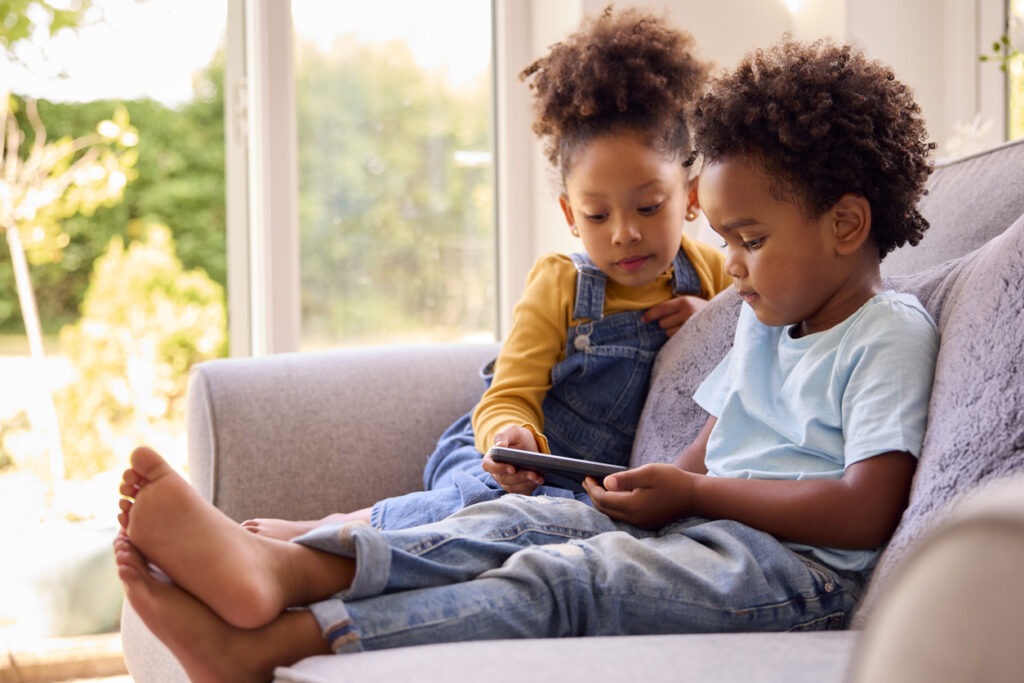 Boy And Girl Playing Handheld Computer Game Sitting On Sofa At Home Together