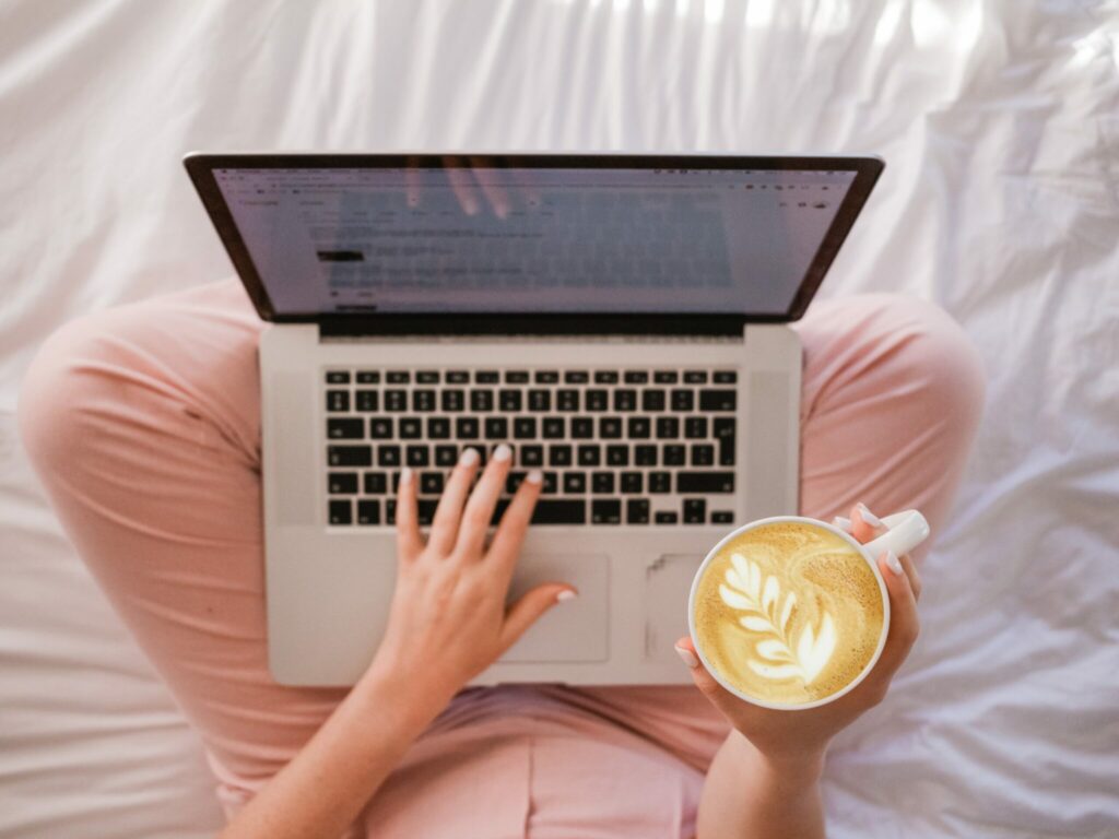 A woman sits on a bed, using a laptop while enjoying a cup of coffee